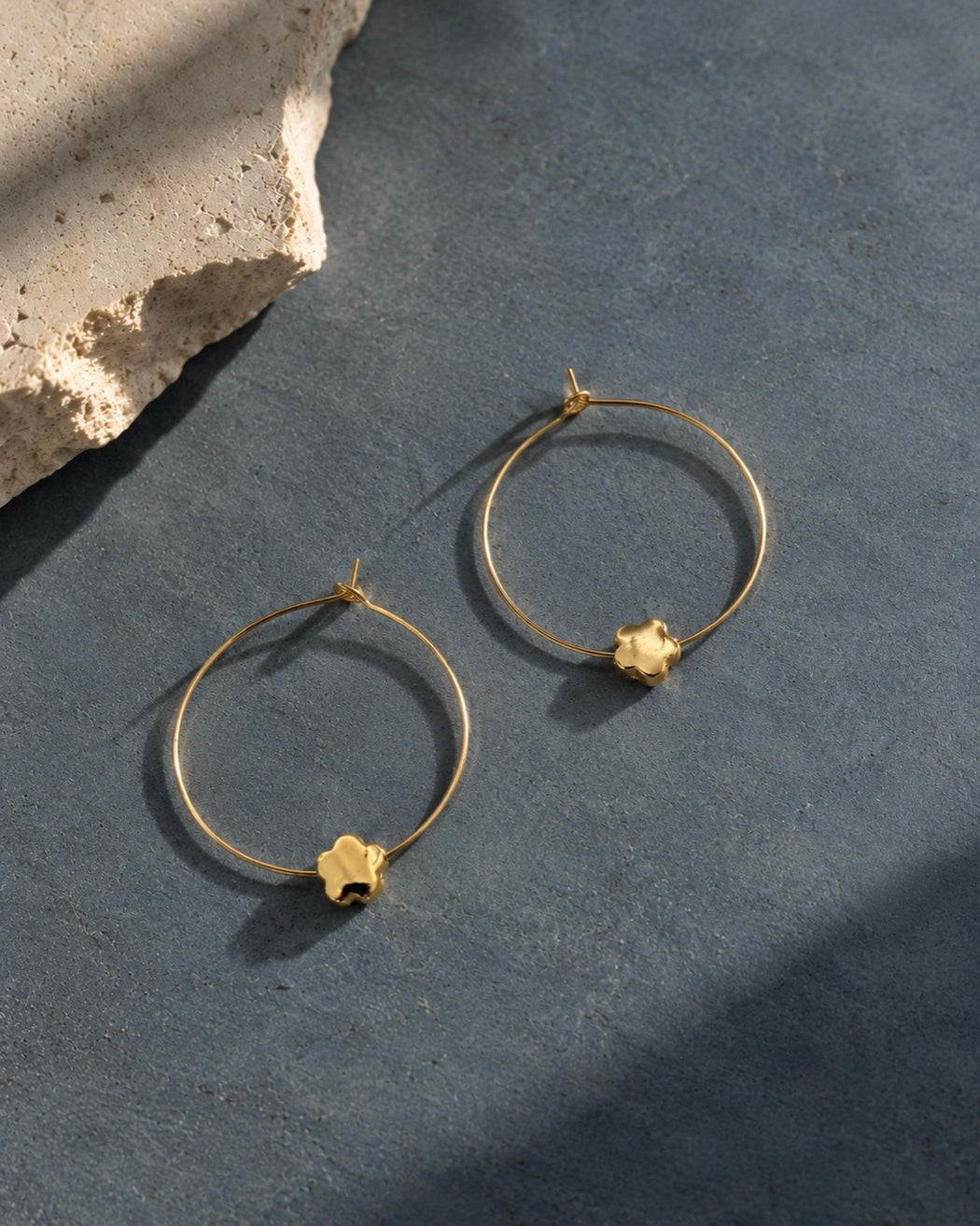 CHLOE gold floral hoop earrings with delicate flower charms on a textured slate blue background.